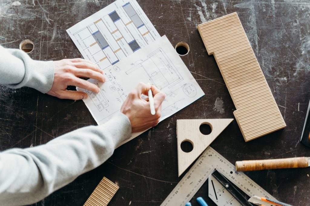 Hands drafting architectural designs on paper with wooden tools in a workshop setting.