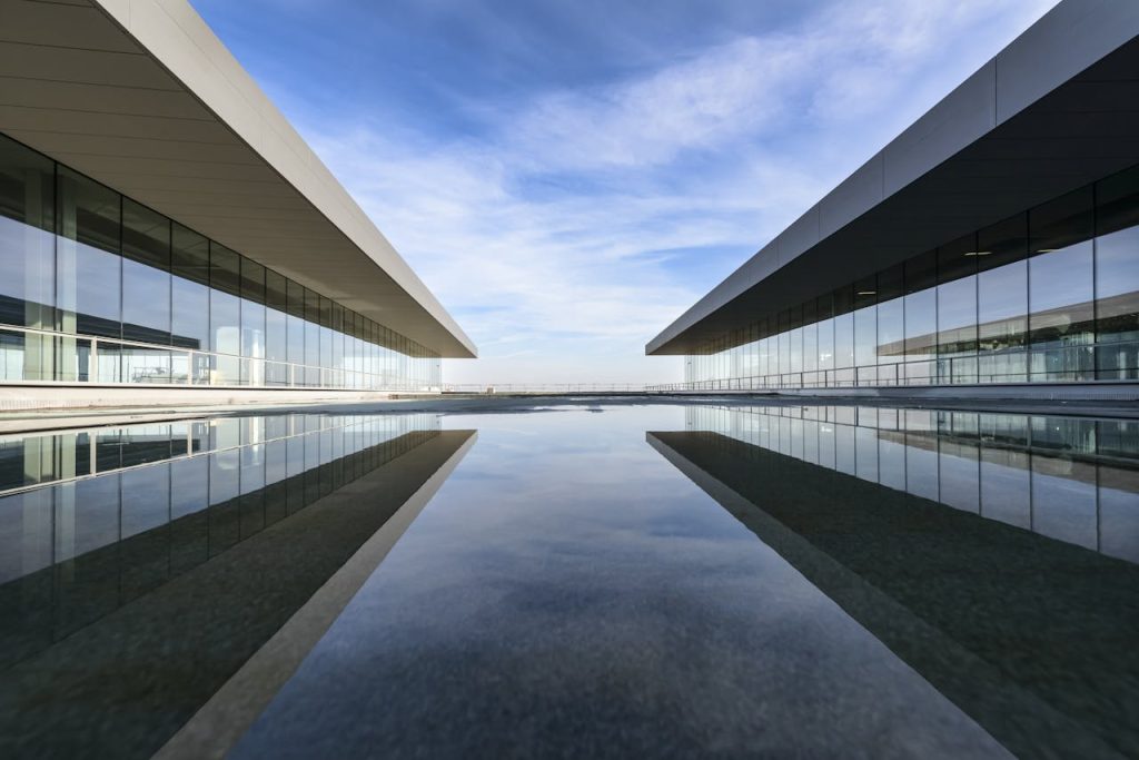 Futuristic glass building mirrored on a still water surface with a cloudy sky.